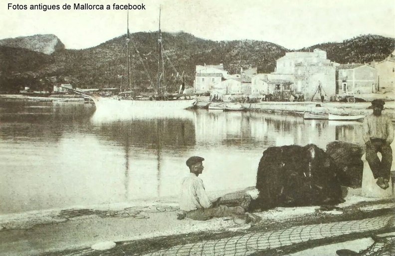 black and white photo of a calm harbor with two fishermen tending nets in the foreground. Sailing boats are docked in the water and modest buildings line the shore against a backdrop of hills. The image depicts a coastal scene in Mallorca from a past era. IMG_17803822575281.jpeg