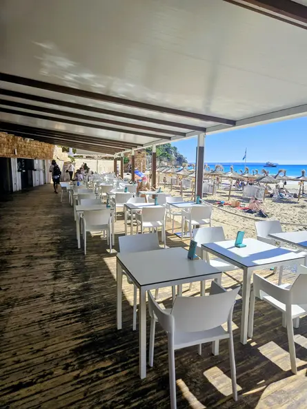 Shaded seaside terrace with white tables beside Cala Major beach and straw umbrellas