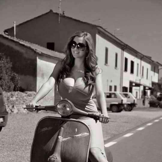 In the black and white photograph, a stylish woman radiates charm atop a classic Vespa scooter. Sporting sunglasses and a casual chic outfit, she embodies the timeless allure of European elegance against a backdrop of quaint buildings. The scene captures a vintage vibe, reminiscent of mid-century dolce vita. FB_IMG_1448844039204.jpg